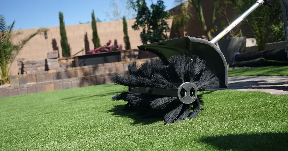 Motorized cleaning brush tool sweeping artificial grass in a landscaped backyard.